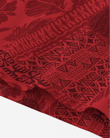 A close-up of folded red fabric with a dark red Polynesian tribal pattern featuring geometric designs, floral motifs, and wave-like stripes.