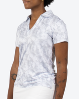 A woman wearing a short-sleeve V-neck polo shirt with a light gray and white abstract botanical pattern, paired with white pants, shown against a plain light gray background.