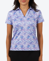 A woman wearing a short-sleeve collared golf shirt with a blue and purple palm leaf pattern on a light lavender background, paired with black bottoms.