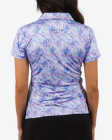 Back view of a woman wearing a short-sleeve collared golf shirt with a blue and purple palm leaf pattern on a white background, featuring the YATTA GOLF logo on the upper back.